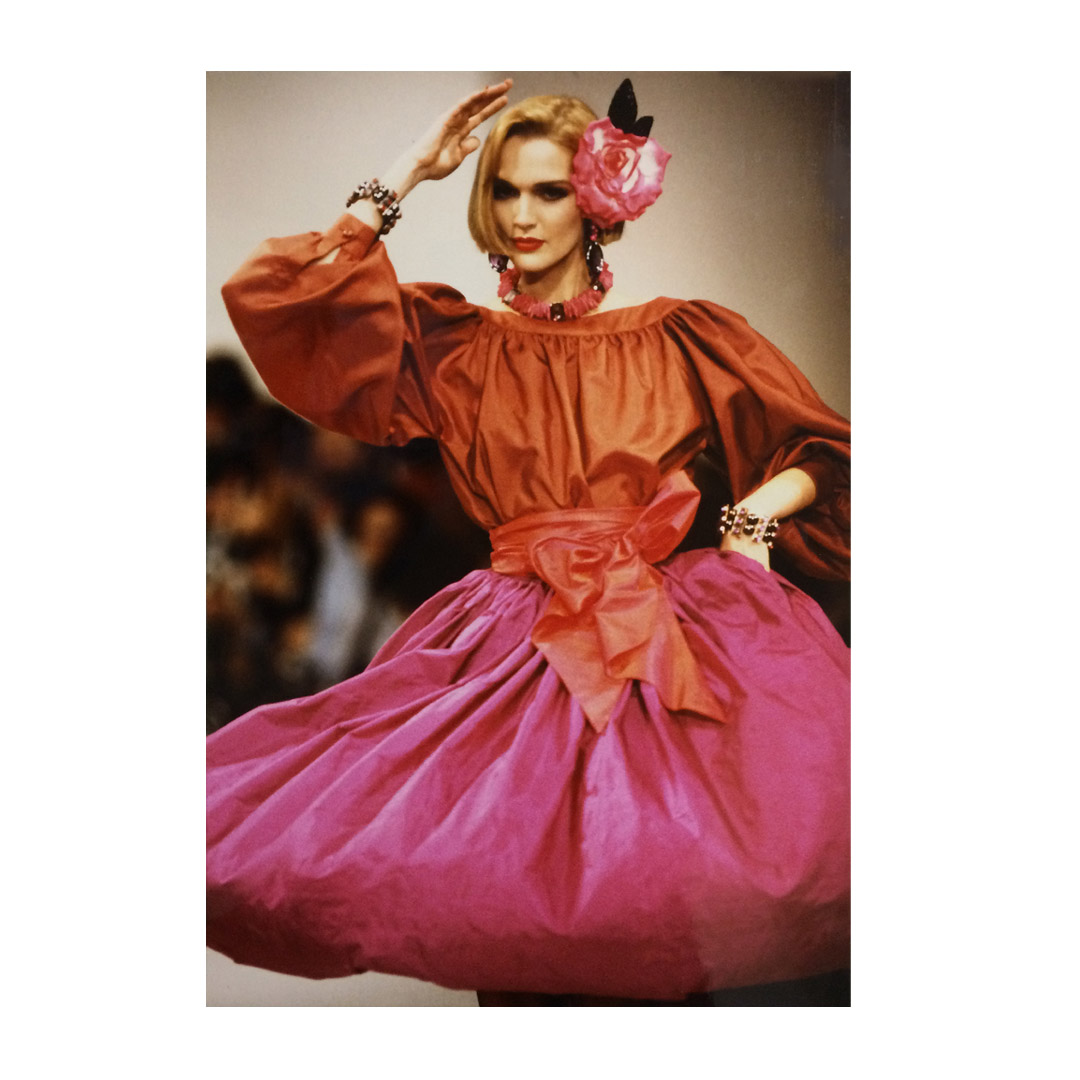 Model on the Paris catwalk in a rust silk blouse and full magenta taffeta skirt with a large bow, a pink rose at the ear — Christian Dior, mid 1980s.