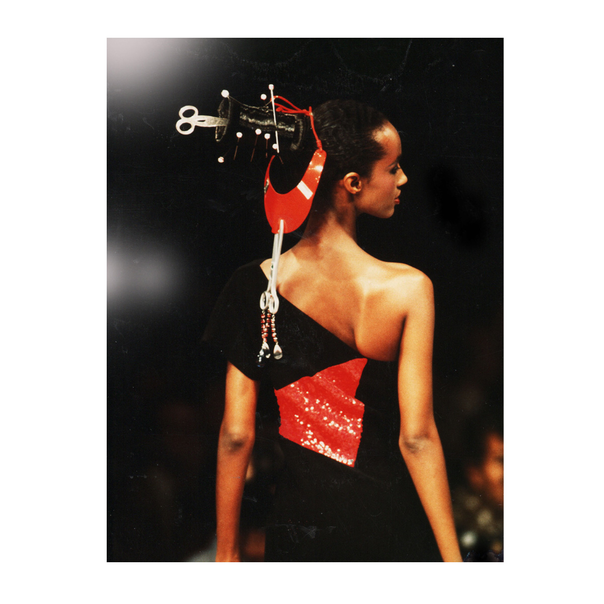 Model photographed from behind on the catwalk in a black one-shoulder gown with a red sequinned panel, wearing a red and black sculptural headpiece with scissors and pins.